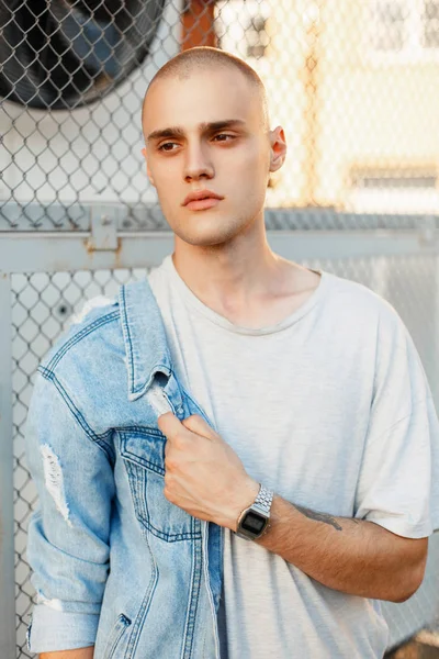 Fashionable portrait of a young brutal man in a stylish denim jacket and a gray shirt near the grid — Stock Photo, Image