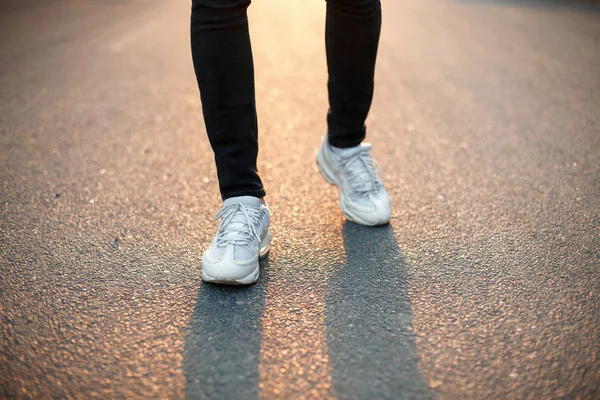 Mannelijke voeten lopen langs een asfaltweg bij zonsondergang. Witte sneakers en zwarte jeans — Stockfoto