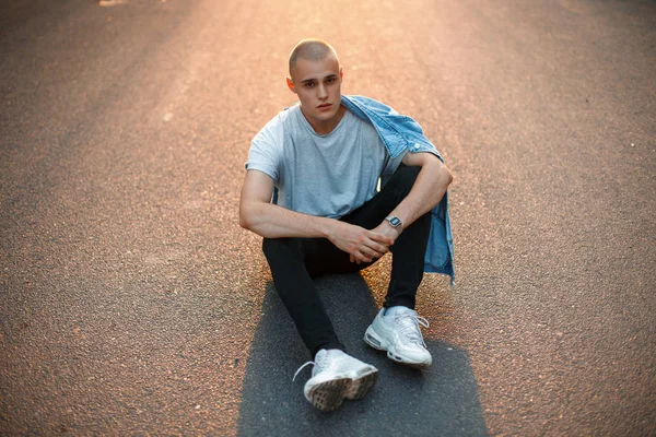 Yakışıklı genç bir gri T-shirt ve şık denim asfalt yolda günbatımında oturur.