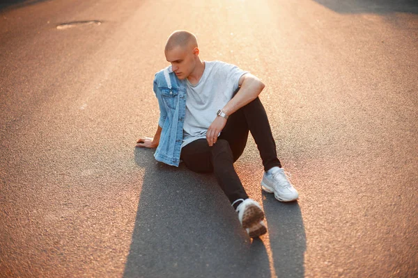 Gri tişört ve kot pantolonla beyaz spor ayakkabı moda yakışıklı acımasız adam asfalt üzerinde günbatımında oturur.