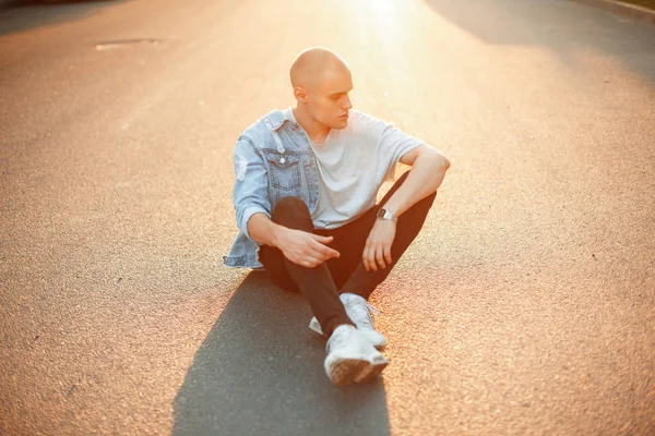 Bir kot ceket gri bir T-shirt ve beyaz spor ayakkabı ile yakışıklı acımasız adam asfalt üzerinde günbatımında oturur.