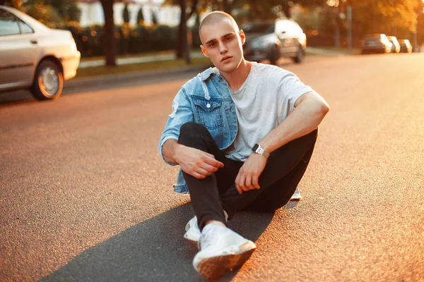Kot elbise ve beyaz spor ayakkabı yakışıklı Amerikan çocuk yolda günbatımında oturur.