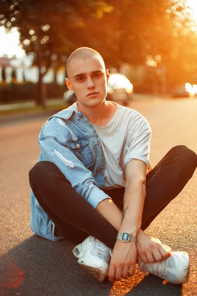 Jeune homme brutal beau dans une veste en denim élégant dans un T-shirt gris et des chaussures blanches se trouve sur l'asphalte au coucher du soleil — Photo