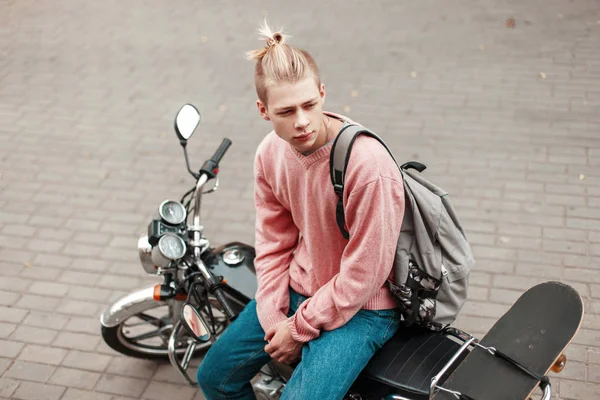 Knappe man met een kapsel in een roze trui met een skateboard en een rugzak zittend op een motorfiets — Stockfoto