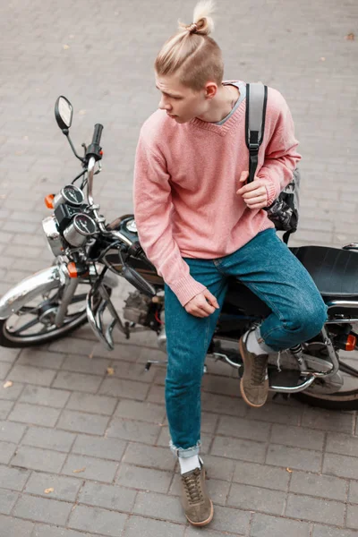 Jovem bonito em um suéter de moda rosa e jeans azul com uma mochila senta-se em uma motocicleta — Fotografia de Stock