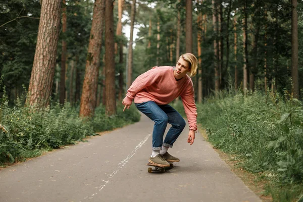 Mladý pohledný mladý muž s účes v růžový svetr brusle na skateboardu v parku — Stock fotografie