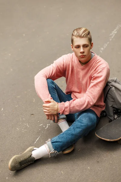 Beau jeune homme dans un pull rose avec une planche à roulettes et un sac à dos assis sur l'asphalte — Photo