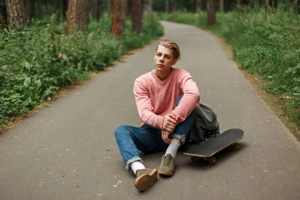 Bonito jovem com um skate em uma camisola rosa e uma mochila senta-se no asfalto — Fotografia de Stock