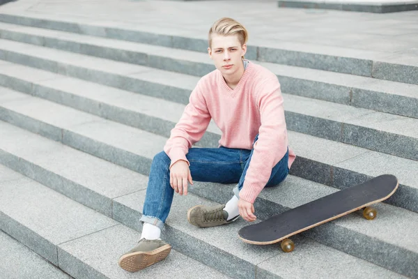 Νέων όμορφος άντρας σε ένα trendy ροζ πουλόβερ με ένα skateboard κάθεται στα σκαλιά — Φωτογραφία Αρχείου