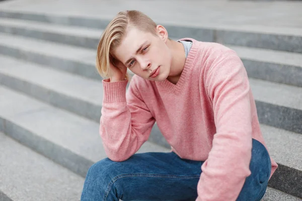 ピンクのおしゃれなセーターで散髪でハンサムな若い男が街の手順に座っています。 — ストック写真