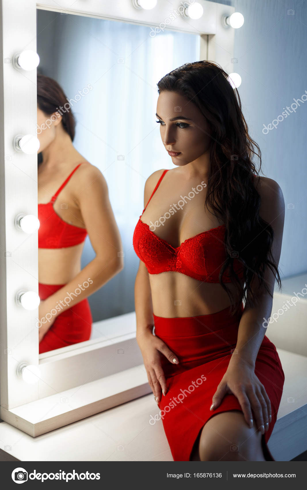 Beautiful young woman with a sexy body in red lingerie and red dress in a bedroom near a vintage mirror with light bulbs Stock Photo by ©alonesdj 165876136 pic