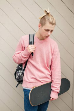 Yakışıklı genç bir pembe kazaklı bir sırt çantası ve ahşap duvar yakınındaki kaykay ile