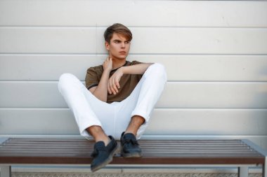 Klasik bir tişört ve siyah ayakkabılar geri kalanı gri parlak duvarının yakınında bir bankta oturan beyaz şık pantolon moda adam