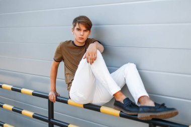 moda yakışıklı bir klasik yeşil T-shirt ve beyaz pantolon bir saç modeli ile modern bir duvara yakın oturur