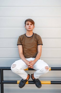 Yakışıklı genç model adam beyaz pantolon ve modern bir duvara yakın oturan siyah ayakkabı ile klasik T-shirt