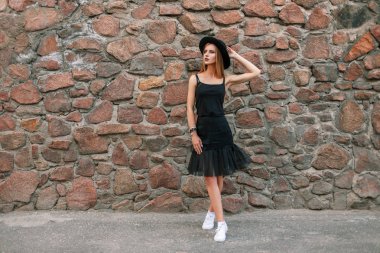 Güzel genç kadın trendy bir şapka ve siyah şık giysiler içinde bir taş duvar yakınındaki beyaz spor ayakkabı