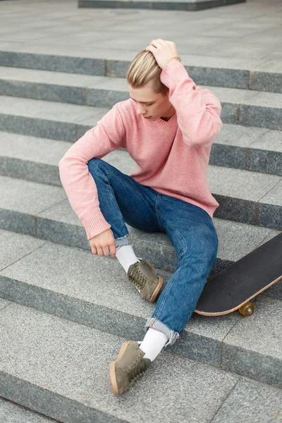 moda yakışıklı genç pembe kazak ve kot bir kaykay ile belgili tanımlık merdiven üstünde oturuyor