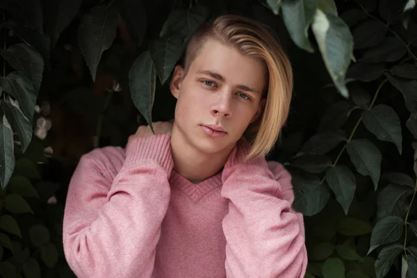 Szép elegáns fiatalember egy frizura, egy rózsaszín divat pulóver közelében egy zöld lombok — Stock Fotó