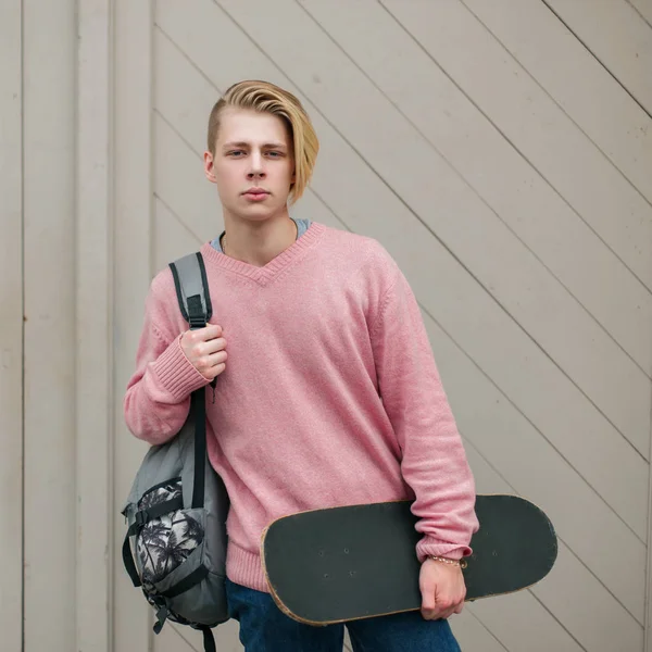 Homem bonito jovem na moda em um suéter rosa com uma mochila e skate perto da parede de madeira — Fotografia de Stock