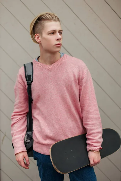 Elegante hombre guapo con un peinado rubio en un suéter rosa con una mochila y un monopatín a la voluntad de una pared de madera — Foto de Stock