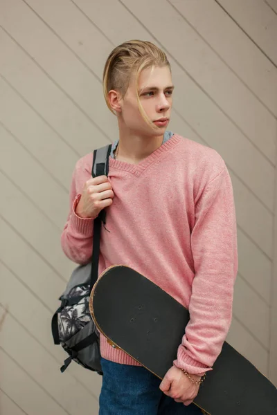 Beau jeune homme avec une coiffure blonde dans un pull rose avec un sac à dos et planche à roulettes près d'un mur en bois — Photo