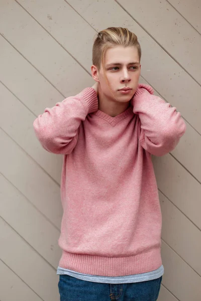 Jeune homme blond à la mode avec une coiffure dans un pull rose près d'un mur en bois — Photo