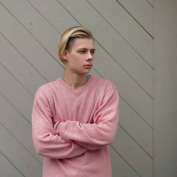 Elegante jovem homem bonito com um penteado loiro em uma camisola rosa perto de uma parede de madeira — Fotografia de Stock