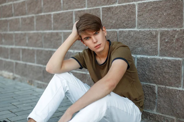 Jovem com um corte de cabelo em uma camiseta clássica está sentado perto da parede em um dia de verão — Fotografia de Stock