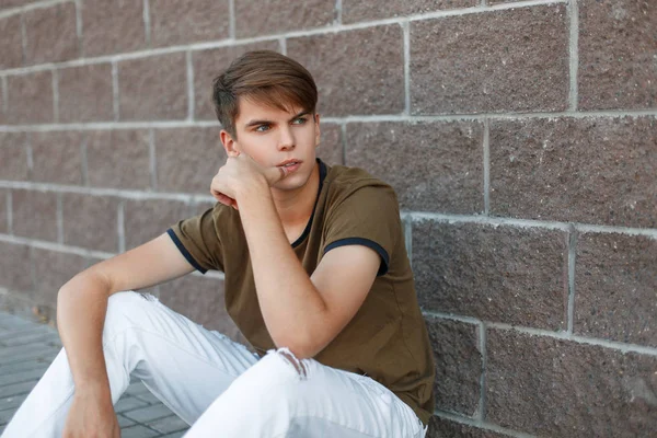 Jeune homme élégant avec des cheveux dans un T-shirt classique assis près du mur un jour d'été — Photo