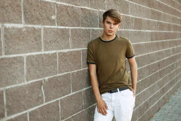 Joven hombre de moda en una camiseta y pantalones blancos cerca de una pared de ladrillo —  Fotos de Stock