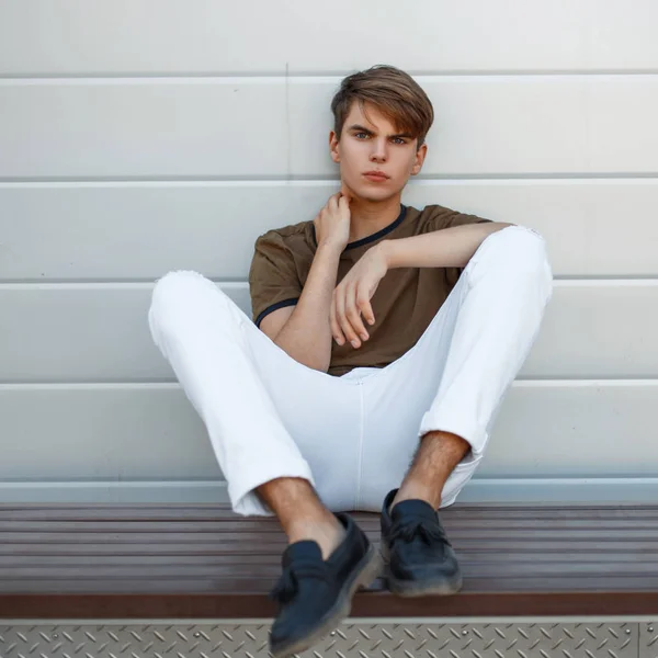 Hombre joven en una camiseta clásica y pantalones blancos con zapatos negros de cuero se sienta cerca de la pared de metal — Foto de Stock