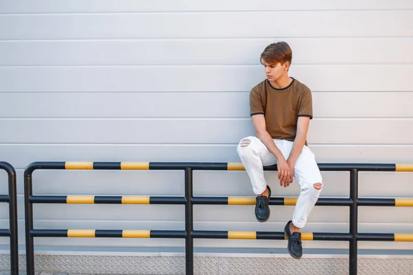 Bonito modelo jovem com um penteado em roupas de moda de verão sentado perto de uma parede cinza brilhante — Fotografia de Stock