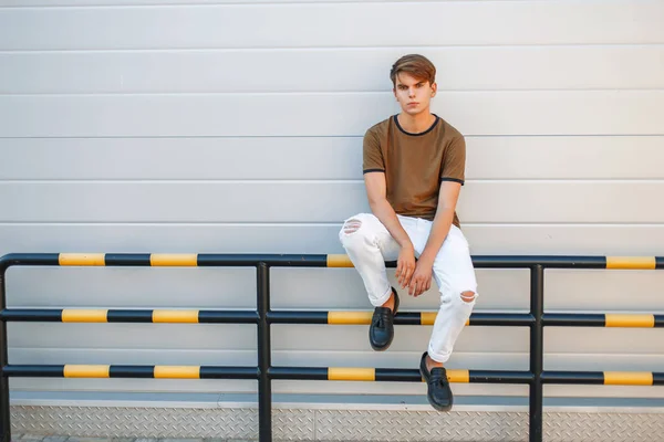 Joven hombre guapo con un peinado en ropa de verano de moda sentado cerca de una pared gris brillante moderno — Foto de Stock