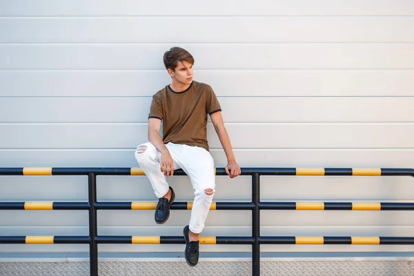 Jovem bonito na moda roupas elegantes senta-se perto de uma parede moderna brilhante — Fotografia de Stock