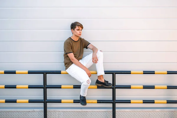 Jovem bonito em uma camiseta clássica na moda e calças brancas com sapatos fica perto de uma parede de metal brilhante — Fotografia de Stock