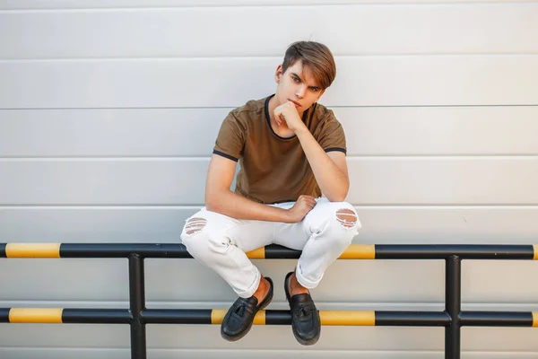 Bonito jovem elegante homem em uma camiseta e calças brancas senta-se perto de uma parede brilhante cinza — Fotografia de Stock