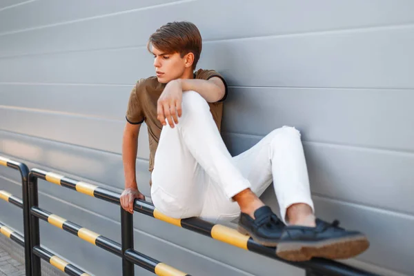 Jovem homem bonito na moda roupas elegantes sentado perto de uma parede de metal — Fotografia de Stock