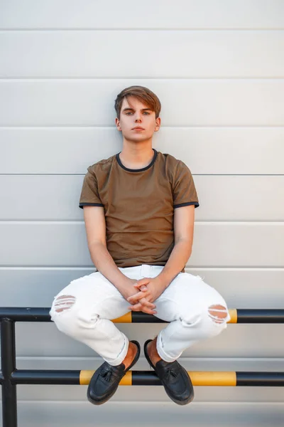 Beau jeune homme modèle en T-shirt classique avec pantalon blanc et chaussures noires assis près d'un mur moderne — Photo