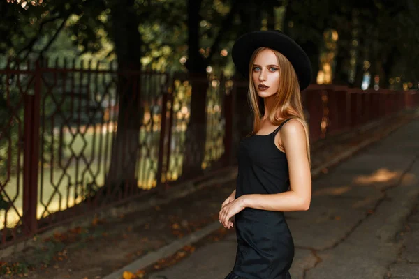 Krásná móda pro mladé ženy v módním černém klobouku a černé oblečení poblíž stromů na ulici — Stock fotografie
