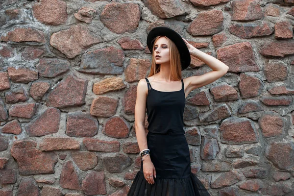 Elegante bella donna in abiti neri alla moda con cappello vicino a un muro di pietra — Foto Stock