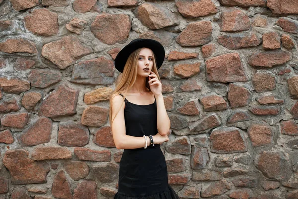 Mooi jong meisje in zwarte mode kleding met een hoed poseren in de buurt van een stenen muur — Stockfoto
