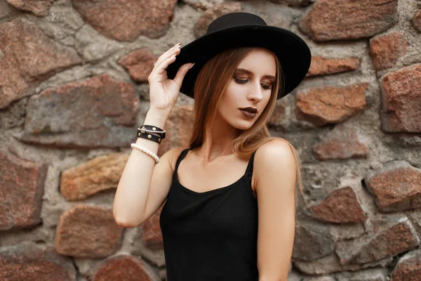 Όμορφο νεαρό μοντέλο γυναίκα στο μαύρο μόδας ρούχα με καπέλο κοντά σε έναν πέτρινο τοίχο — Φωτογραφία Αρχείου