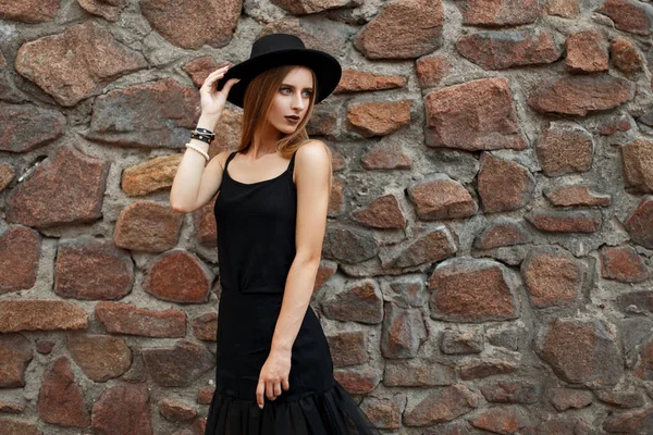 Όμορφη νεαρή γυναίκα σε κομψό μαύρο καπέλο και μοντέρνα ρούχα κοντά σε ένα παλιό πέτρινο τοίχο — Φωτογραφία Αρχείου