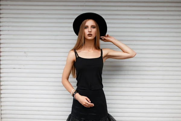 Bella donna elegante in abiti di moda neri con un cappello vicino al muro bianco — Foto Stock