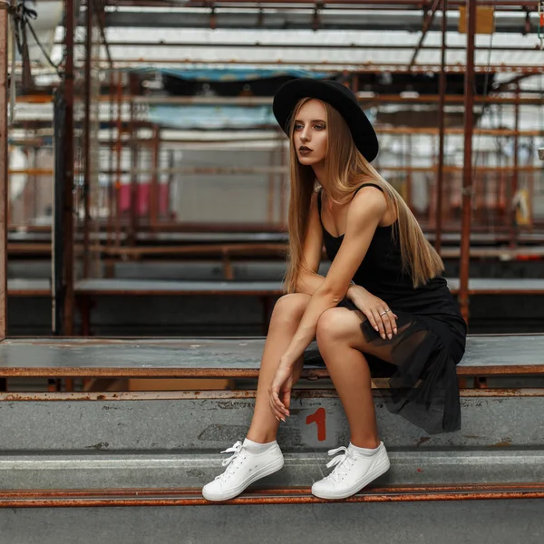 Mooie stijlvolle vrouw met een trendy hoed en een zwarte rok met witte schoenen zit op de straat — Stockfoto