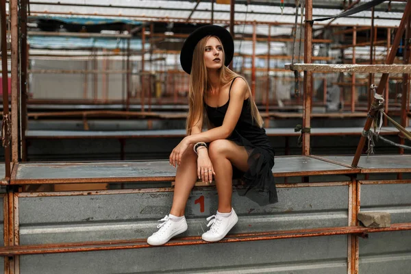 Beautiful young model woman in stylish black clothes with a trendy hat and white shoes sitting on a table — Stock Photo, Image