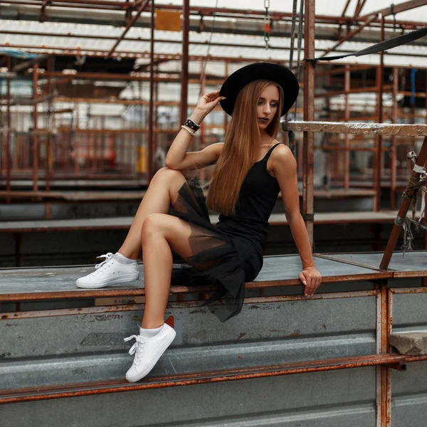 Mooie modieuze vrouw in een zwarte hoed en een stijlvolle rok in witte schoenen zit op een metalen tafel — Stockfoto