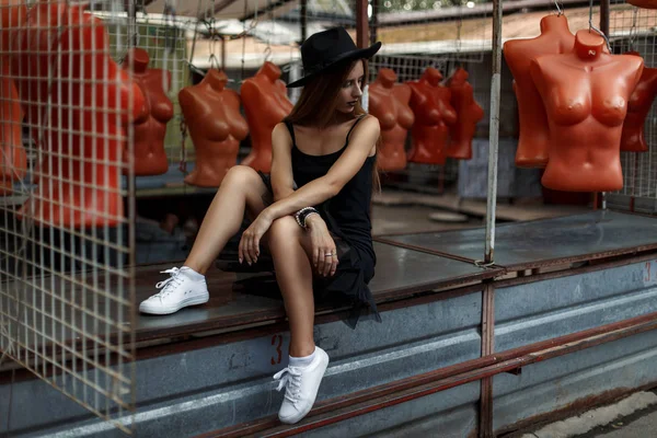 Mooie modieuze meisje in zwarte stijlvolle kleding met een hoed zit in de buurt van de mannequins — Stockfoto