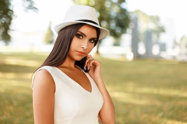 Mooie jonge vrouw in een trendy witte jurk en een stijlvolle hoed buiten in het park — Stockfoto