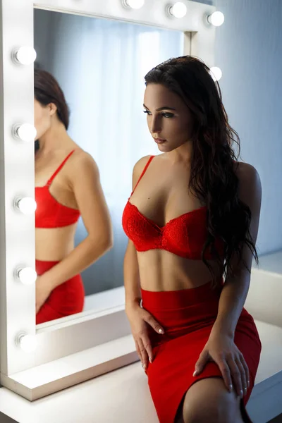 Beautiful young woman with a sexy body in red lingerie and red dress in a bedroom near a vintage mirror with light bulbs — Stock Photo, Image